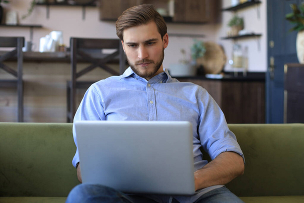 Concentrated young freelancer businessman sitting on sofa with laptop, working remotely online at home - Photo, image