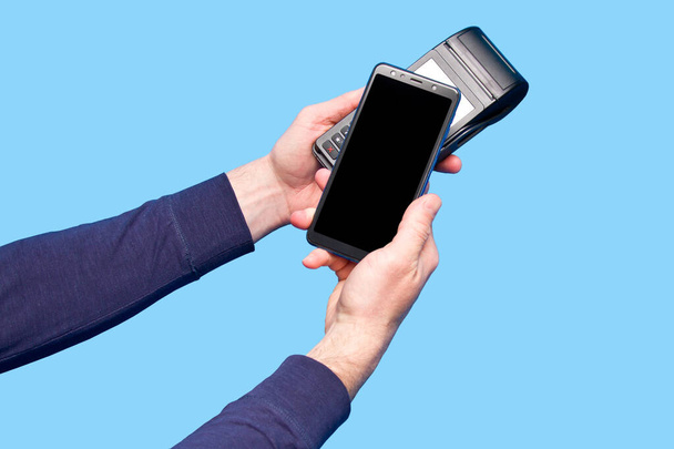 Male hand using contactless payment terminal with smartphone on blue background. - Photo, Image