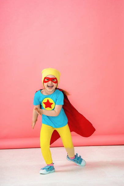 a little girl in a superhero costume in a red Cape and hat shows, runs forward. The baby is laughing merrily and posing with pleasure in the Studio on a pink background. - Foto, Bild