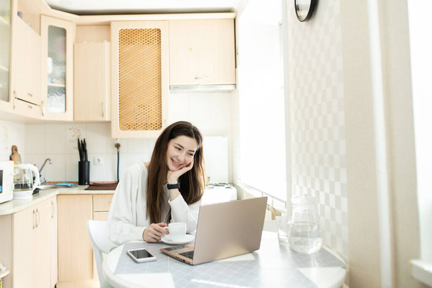 Usmívající se dívka čte doma e-mailovou kuchyni. Podnikatelka pije kávu a pracuje s laptopem. Talentovaný manažer dálkově pracuje v e-mailu pro psaní notebooků - Fotografie, Obrázek