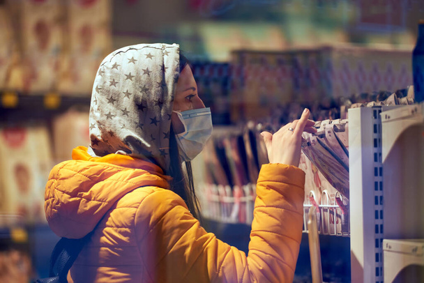 Ein Mädchen mit medizinischer Maske, Kapuze und Aktentasche wählt einen Artikel im Geschäft. Dame in orangefarbener Jacke. war des Covid-19 müde. Der Hintergrund ist unklar. - Foto, Bild