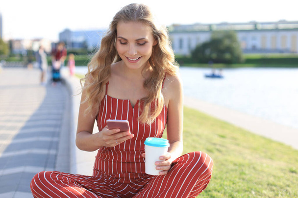 Portrait of pretty young woman sitting on riverbank with legs crossed during summer day, using smartphone. - Fotoğraf, Görsel