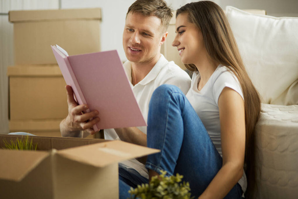 Cute couple unpacking cardboard boxes in their new home, sitting on the floor and looking at a family album - Photo, image