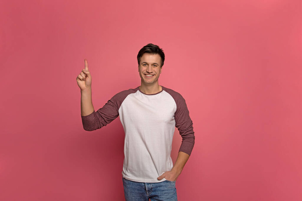 Advertisement. Close-up photo of a European man in casual clothes, who is pointing upwards with his right index finger and smiling, while looking in the camera. - Valokuva, kuva