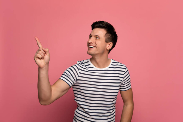 My favourite view. Close-up photo of cheerful young man in a striped t-shirt, who is smiling while pointing to the left upper corner. - Photo, Image