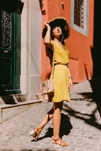 Yazlık elbiseli ve çantalı hasır şapkalı çekici bir kadın. Esmer, uzun saçlı, sarı yazlık elbiseli, trav şapkalı ve saman çantalı bir kadının portresi eski şehir sokaklarında yürüyor.. - Fotoğraf, Görsel