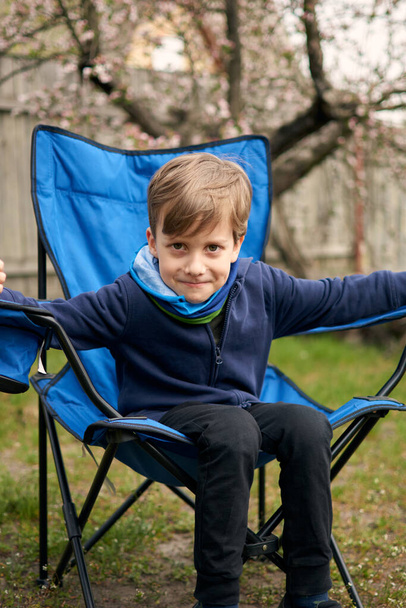 Niño de 6 años sentado en una silla de camping solo relajante outdors. Disfruta de la naturaleza. Abuela casa de campo
. - Foto, Imagen