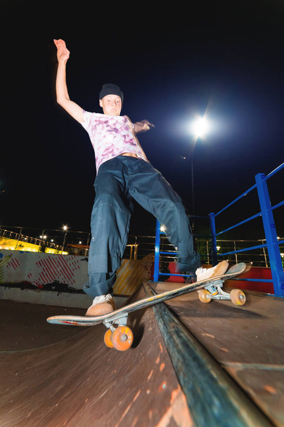 A young skater does the trick of sliding at night in a skatepark amid the burning lights of lighting poles. The concept of night leisure and youth culture at night - Photo, Image