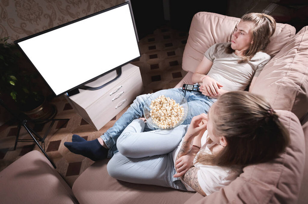 Atrakcyjna młoda, długowłosa para przytulająca się na kanapie w domu w izolacji oglądając telewizję. Koncepcja spędzania czasu z bliskimi podczas pandemii. - Zdjęcie, obraz