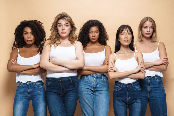 Image of sassy multinational women in blue jeans posing with hands crossed isolated over beige background - Valokuva, kuva