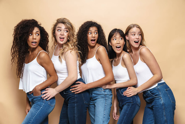 Image of beautiful multinational women in blue jeans expressing surprise and looking aside isolated over beige background - Photo, image