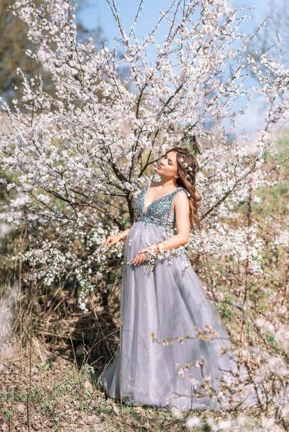 Schöne schwangere Frau in einem eleganten Kleid in einem blühenden Frühlingsgarten - Foto, Bild