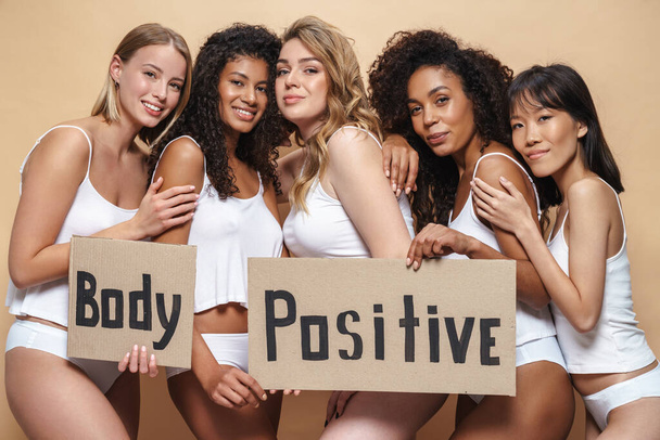 Imagen de mujeres multinacionales alegres en ropa interior abrazando y sosteniendo pancartas aisladas sobre fondo beige
 - Foto, imagen