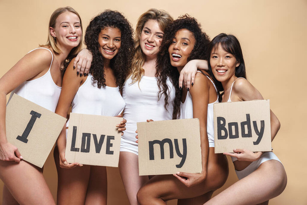 Image of cheerful multinational women in underclothes hugging and holding placards isolated over beige background - Foto, afbeelding