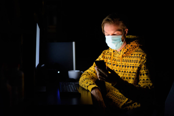 夜遅くに隔離中に自宅から働くコロナウイルス感染から保護するためのマスクを持つ若い男の肖像画 - 写真・画像
