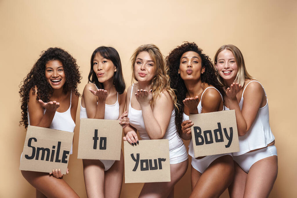 Image of pleased multinational women in underclothes holding placards and sending air kisses isolated over beige background - 写真・画像