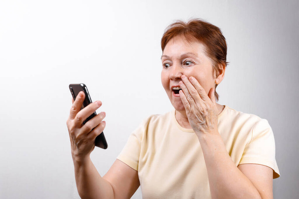 старша жінка дивиться здивовано по телефону на білому тлі в легкій футболці. місце для тексту. ізольовані
 - Фото, зображення