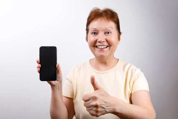 Senior woman shows a phone on a white background in a light T-shirt. place for text. isolated - Photo, image