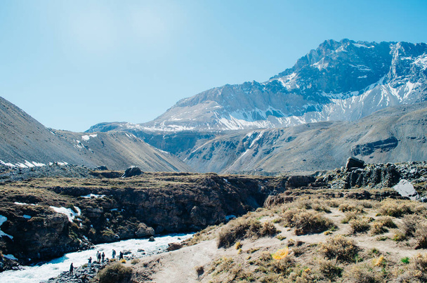 Rocky trekking path of the Andes Mountains with a group of people near a river and with a huge mountain partially covered in snow in the background with clear blue skies and a sun flare. - Foto, Bild