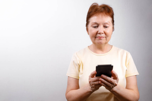 Az idősebb nő fehér alapon nézi a telefont egy könnyű pólóban. SMS-ezés helye. izolált - Fotó, kép