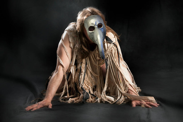Half-naked man with long hair in medieval rags and a plague mask on a black background - Photo, Image