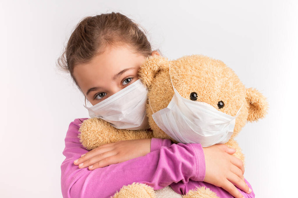 Little Russian Girl In Medical Mask Protection From Influenza Holds In Her Embrace Soft Medium Toy Bear In Mask On White Background Close-Up. - Φωτογραφία, εικόνα