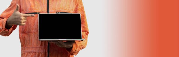 Le chef mécanicien dans un uniforme orange tenant écran vierge ordinateur portable. Debout avec son doigt pointant vers l'ordinateur. Portrait avec lumière de studio
. - Photo, image