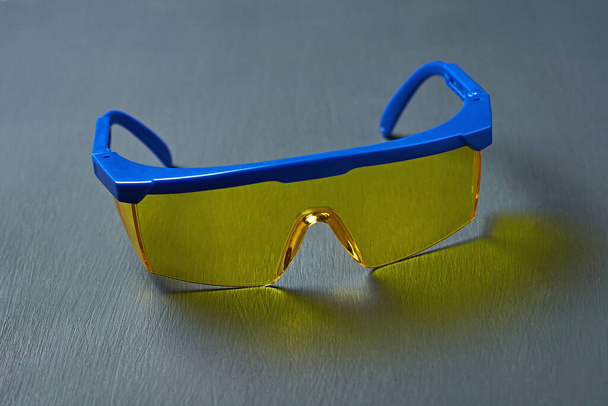Skibril of moderne veiligheidsbril voor arbeider op de bouw, lasser, tandarts, turner en andere met geel glas en blauw frame ligt op donker betonnen bureau. Close-up - Foto, afbeelding