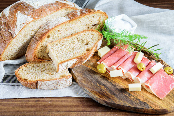 Tahta kesme tahtasında dilimlenmiş taze jamon, peynir ve zeytin. Mutfak peçetesinde ev yapımı integral ekmek ve tuz. Doğal ev yapımı sağlıklı gıda konsepti.  - Fotoğraf, Görsel
