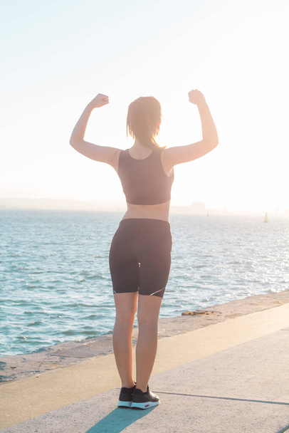 Visão traseira da menina de esportes com os braços levantados de pé ao ar livre perto do mar sentindo forte
 - Foto, Imagem