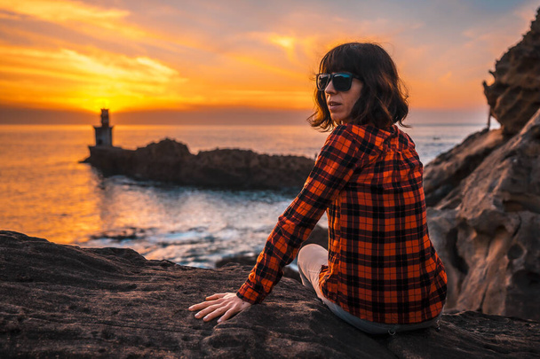 A young girl sitting with sunglasses in the orange sunset of the Pasajes San Juan lighthouse, near San Sebastian, Gipuzkoa. Basque Country - Valokuva, kuva