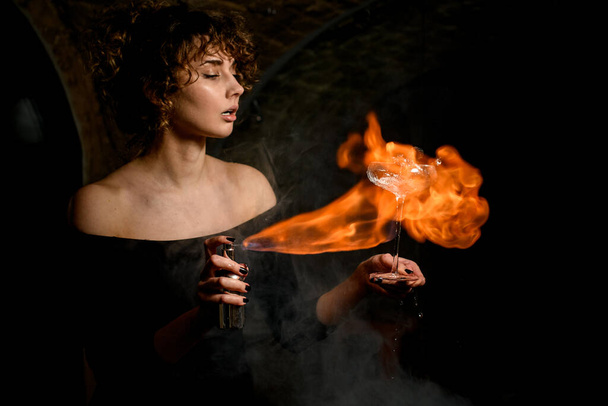 attractive woman holds glass with splashing drink, sprinkles and sets fire to it. - Photo, Image