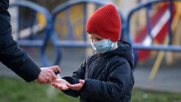 Απολυμαντικό χεριών σε δημόσιο χώρο. Μια μητέρα με ιατρική μάσκα πιτσιλάει το προϊόν στα χέρια του παιδιού. Η έννοια της προστασίας κατά των λοιμώξεων από τον ιό Coronavirus covid-19 - Πλάνα, βίντεο