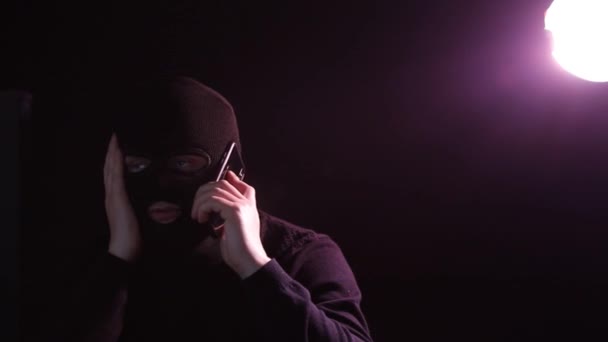 Een man in een bivakmuts in een donkere kamer pratend op een smartphone. - Video