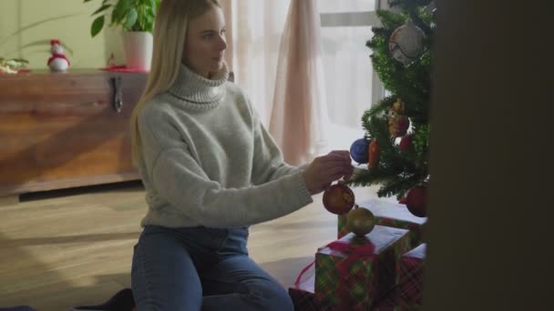 Beautiful woman decorating christmas tree with toys by the window - Filmati, video
