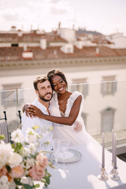 Interracial wedding couple. Destination fine-art wedding in Florence, Italy. African-American bride and Caucasian groom are sitting at the rooftop wedding dinner table overlooking the city. - Photo, Image