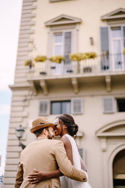 Esküvő Firenzében, Olaszországban. Multiracionális házaspár. Afrikai-amerikai menyasszony fehér ruhában, hosszú fátyollal és csokorral, és fehér vőlegény homokos dzsekiben és szalmakalapban.. - Fotó, kép