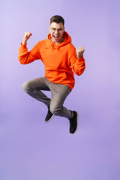 Image of happy emotional young man jumping isolated over purple background wearing eyeglasses showing winner gesture. - Foto, imagen