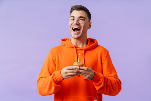 Photo of cheerful optimistic happy young man posing isolated over purple background holding birthday cupcake with candle. - Photo, Image