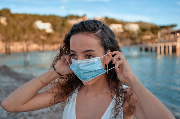 Πορτρέτο της νεαρής γυναίκας που στέκεται στην παραλία. Απολαμβάνοντας μια μέρα στην παραλία με μια ιατρική μάσκα. - Φωτογραφία, εικόνα