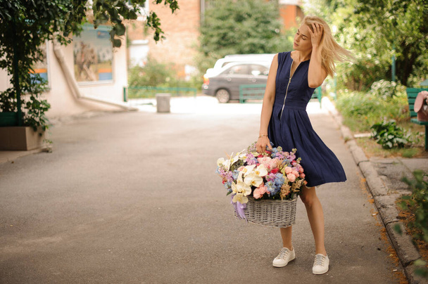 young girl in blue dress holds basket with flowers in one hand and straightens her hair with other hand - Fotó, kép