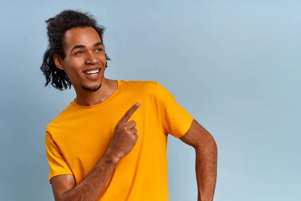 Cute african african smiling fashion male in a yellow t-shirt points his finger right up, isolated on a blue background. Concept of services and products in online stores for youth. Copy space - Foto, Imagem
