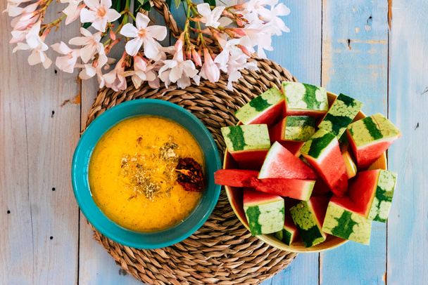 Felett színes élelmiszer kilátás sárga zöldség leves és piros friss görögdinnye gyümölcs egy kék sárga asztal virág dekoráció - Fotó, kép
