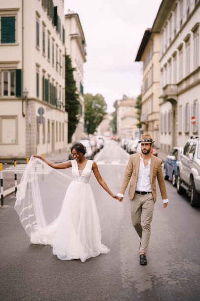 Svatební dvojice smíšených ras. Svatba ve Florencii, Itálie. Africko-americká nevěsta v bílých šatech a kavkazský ženich v klobouku kráčejí po cestě mezi auty. - Fotografie, Obrázek