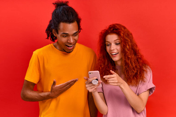 Young mixed race couple african american man and charming caucasian woman watching online store with furniture using smartphone and Internet on red background. Relocation concept. Advertising space - Foto, Imagem
