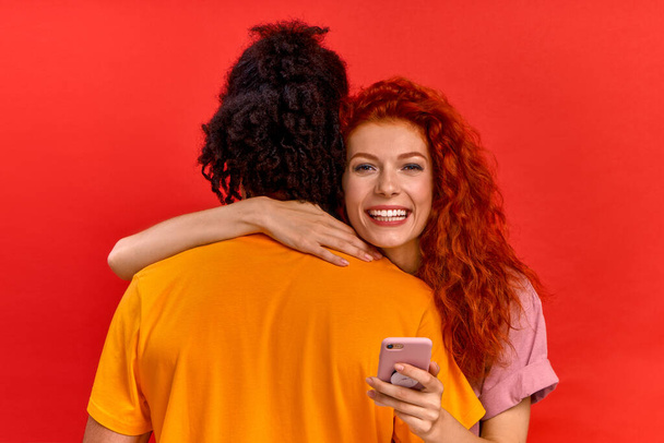 Cute young happy red haired caucasian girl hugs her african american boyfriend and looks at modern smart phone. Concept of online dating site and applications for lovers. Advertising space - Fotografie, Obrázek