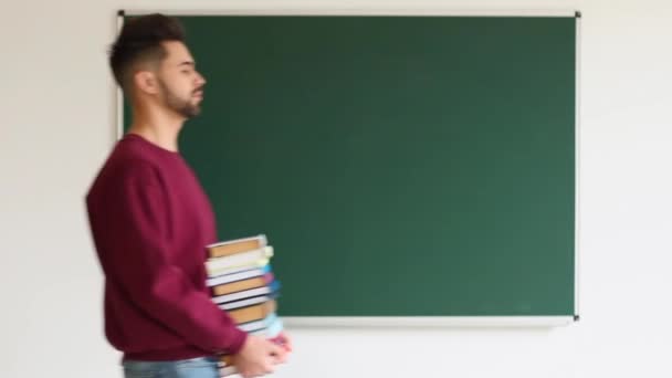 Κουρασμένος μαθητής με πολλά βιβλία που περνούν από μαυροπίνακα στην τάξη - Πλάνα, βίντεο