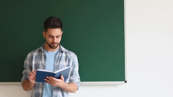mężczyzna student czytanie książki w pobliżu tablicy w klasie - Materiał filmowy, wideo