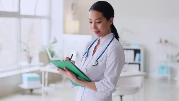 Female Asian doctor writing on clipboard in clinic - Footage, Video