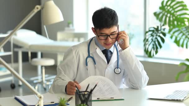 Médico asiático hablando por teléfono en la mesa en la clínica
 - Imágenes, Vídeo